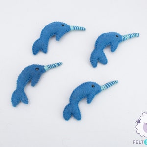May include: Four blue felt narwhal ornaments with white and blue striped horns. The narwhals are arranged in a circle on a white background.