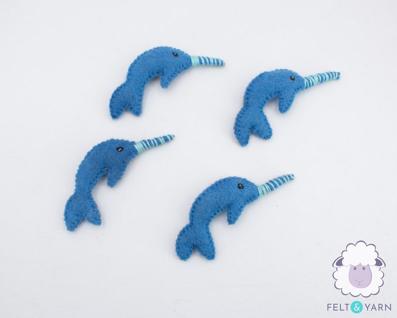 May include: Four blue felt narwhal ornaments with white and blue striped horns. The narwhals are arranged in a circle on a white background.