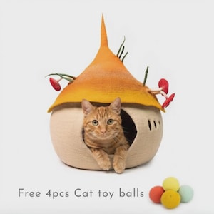 May include: A beige felt cat cave with a pointed orange roof, decorated with small green stems and red mushroom accents. An orange tabby cat sits inside the opening. Four small, colorful felt balls are placed in front of the cat cave.