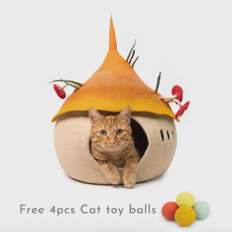 May include: A beige felt cat cave with a pointed orange roof, decorated with small green stems and red mushroom accents. An orange tabby cat sits inside the opening. Four small, colorful felt balls are placed in front of the cat cave.