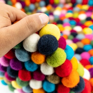Pu&ograve; includere: Un tappeto colorato fatto di palline di feltro in varie tonalit&agrave; di rosso, blu, verde, giallo, bianco e viola. Le palline sono disposte in un motivo circolare, creando una superficie unica e testurizzata.