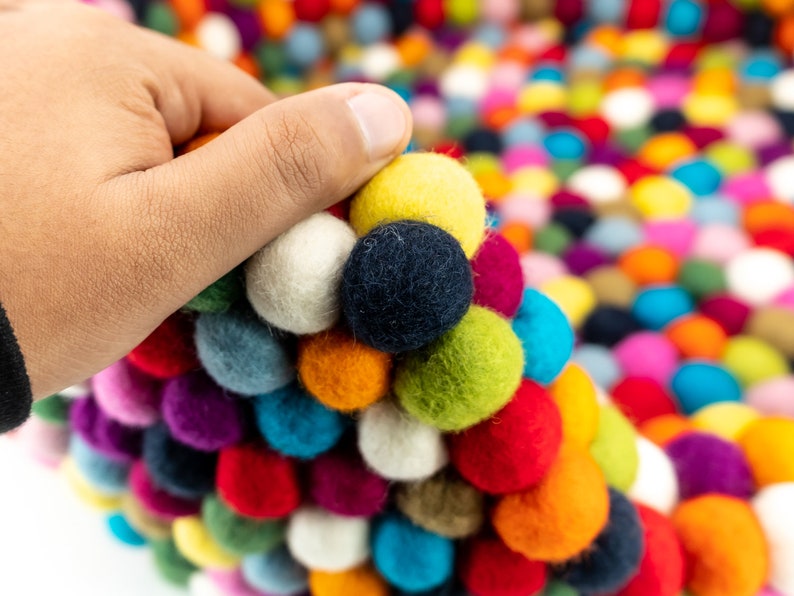 Pu&ograve; includere: Un tappeto colorato fatto di palline di feltro in varie tonalit&agrave; di rosso, blu, verde, giallo, bianco e viola. Le palline sono disposte in un motivo circolare, creando una superficie unica e testurizzata.