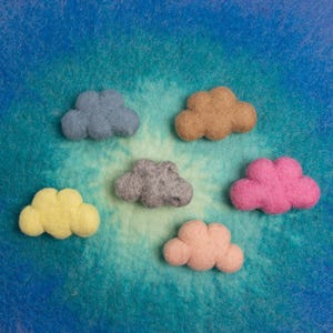 Może przedstawiać: Sześć chmur z filcu w różnych kolorach, w tym różowym, żółtym, szarym, niebieskim i brązowym, ułożonych na niebieskim i zielonym tle.