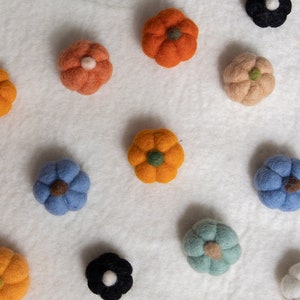 May include: A collection of felt pumpkins in various colors, including orange, blue, green, and white. The pumpkins are arranged on a white background.
