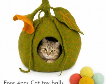 May include: A green felted wool cat cave shaped like a fruit with a stem, a large leaf, and an orange flower accent. A brown tabby cat sits inside the circular opening. Four small, round, multi-colored felt balls are placed nearby.