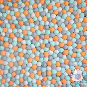 May include: A colorful felt ball rug with a repeating pattern of orange, gray, light blue, and white spheres. The rug is made of many small, round felt balls, creating a textured surface. The rug is a decorative item.
