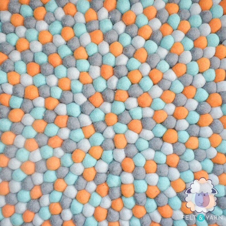 May include: A colorful felt ball rug with a repeating pattern of orange, gray, light blue, and white spheres. The rug is made of many small, round felt balls, creating a textured surface. The rug is a decorative item.