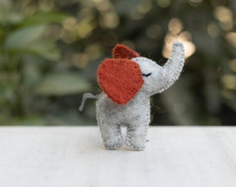 May include: A small gray felt elephant toy with red felt ears. The elephant is standing on a white surface.