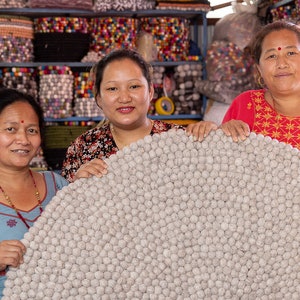 May include: Three women are holding a large, round, gray, wool rug made of pom-poms. The rug is in front of a shelf with colorful, round, felt balls.