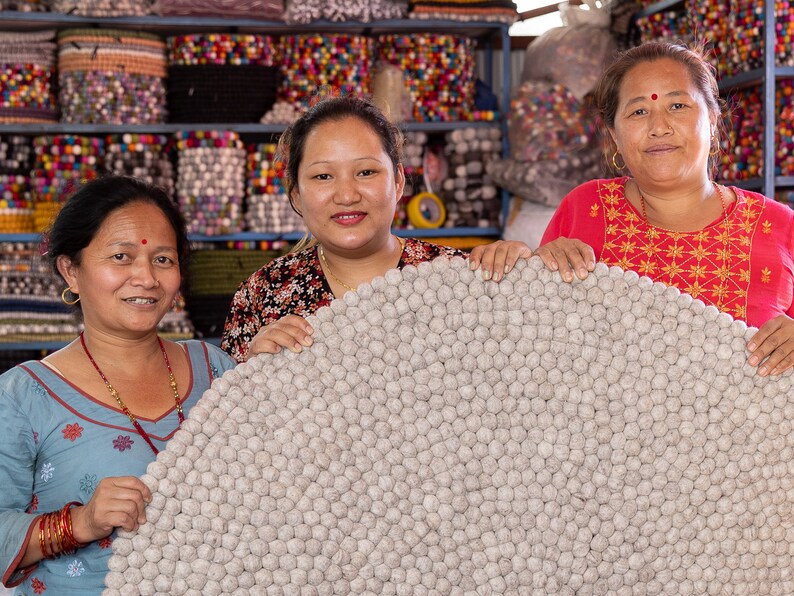 May include: Three women are holding a large, round, gray, wool rug made of pom-poms. The rug is in front of a shelf with colorful, round, felt balls.