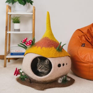 May include: A felted cat cave shaped like a mushroom house with a beige base, a yellow and orange pointed roof, and red mushroom accents. A tabby cat sits inside the circular opening. The structure features small green felted grass details.