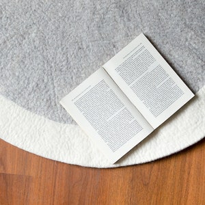 May include: A white and gray round rug with a book lying on top of it. The rug is on a wooden floor.