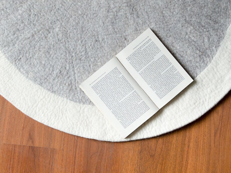 May include: A white and gray round rug with a book lying on top of it. The rug is on a wooden floor.