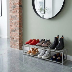 May include: A silver metal shoe rack with two tiers, displaying a variety of footwear. The shoes include red loafers, sneakers, black ankle boots, sandals, and high-top sneakers. The rack is positioned against a light green wall.