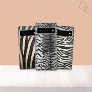 May include: Three phone cases with animal print designs. The cases feature a zebra print, a tiger print, and another zebra print. The cases are standing on a white surface with a peach-coloured background.