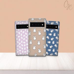 May include: Three phone cases with a repeating bunny pattern. The cases are in pastel colors: lavender, beige, and blue. Each case has a camera cutout and a white bunny face design.