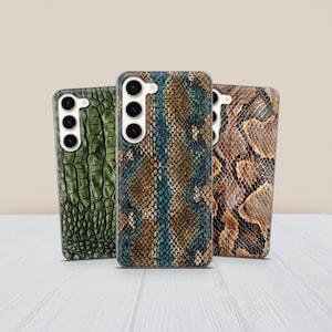 Pode incluir: Três capas de telemóvel com padrões de pele de réptil. As capas apresentam uma textura de pele de crocodilo verde, um design de pele de cobra azul e castanho e um padrão de pele de cobra castanha. Cada capa tem um recorte para a câmara.