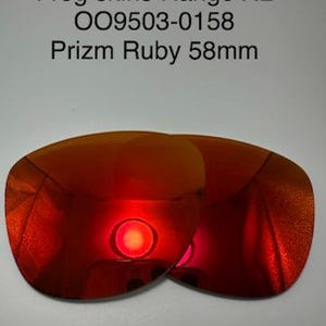 Pode incluir: Duas lentes de substituição para óculos de sol Prizm Ruby com acabamento vermelho refletivo. O texto diz: Frog skins Range XL, OO9503-0158, Prizm Ruby 58mm. Estas lentes apresentam uma forma curva e um tom vermelho metálico vibrante.