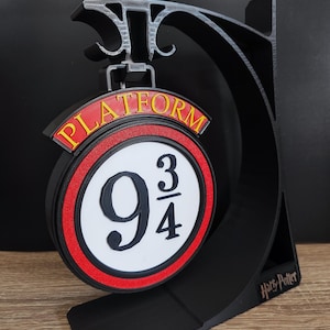 Peut inclure: Panneau noir et gris imprimé en 3D pour la plateforme 9 3/4. Le panneau présente un design circulaire avec les chiffres "9 3/4" et le mot "PLATFORM" en lettres jaunes. Le panneau est monté sur un support noir.