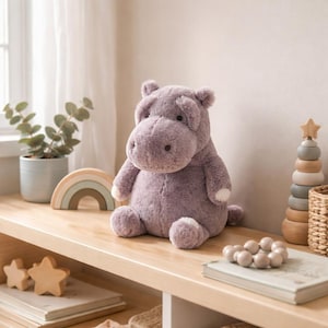 Puede incluir: Un peluche de hipopótamo lavanda con detalles blancos en los pies y las orejas está colocado en una estantería de madera. Otros elementos decorativos incluyen un arcoíris de madera, una planta en una maceta azul y un juguete apilable de madera.