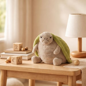 Puede incluir: Un peluche de tortuga con un caparazón verde claro y una expresión de mal humor está sentado en una mesa de madera. La tortuga es un animal de peluche con un cuerpo beige y brazos y piernas pequeños. Una lámpara de madera y bloques también están en la mesa.