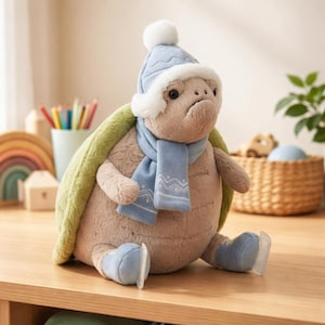 Puede incluir: Un peluche de tortuga con un gorro de invierno azul claro con un pompón blanco, una bufanda a juego y patines de hielo. La tortuga tiene un caparazón verde y está sentada sobre una superficie de madera.