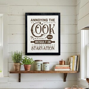 May include: A kitchen scene featuring a framed sign with the text "Annoying the Cook Will Result in Starvation." A wooden shelf holds potted herbs, jars, and books. A copper pot sits on a gas stove, and a white sink is visible.