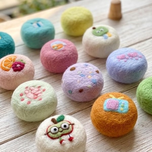 May include: A collection of round, felted wool balls in various colors including yellow, pink, blue, orange, and white. Each ball features a small, needle-felted design such as a television, fruit, hearts, or a cartoon character with glasses.