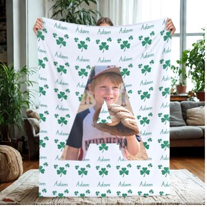 Puede incluir: Manta de forro polar blanca con un patrón de tréboles verdes y el nombre "Adam" repetido. Un recorte triangular revela una foto de un niño con uniforme de béisbol sosteniendo una pelota y un guante. La palabra "MINHENS" es visible en el uniforme.