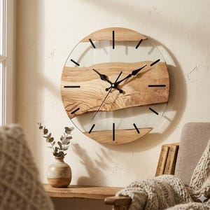 Puede incluir: Un reloj de pared redondo con un diseño de madera natural y vidrio. La esfera del reloj presenta un centro de veta de madera de color claro con manecillas y marcadores de hora negros. El reloj está montado en una pared de color claro.