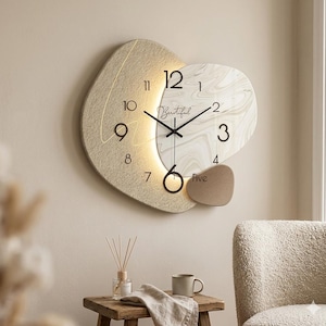 May include: A modern wall clock with a unique design. The clock features a beige, abstract shape with a marble-like face and gold-colored hands. The word "Beautiful" is written on the clock face. The clock is illuminated with a soft light.