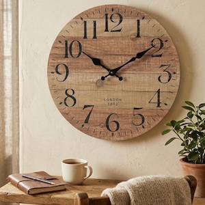Puede incluir: Un reloj de pared redondo de madera con números y manecillas negras. La esfera del reloj tiene un patrón de vetas de madera marrón claro y el texto "LONDON 1892". El reloj está colgado en una pared beige.