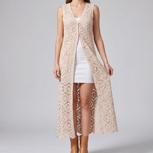 Può includere: Gilet lungo senza maniche in crochet beige con scollo a V e chiusura con bottoni. Il gilet presenta un design aperto e fluido con intricati motivi floreali. L'indumento arriva al ginocchio ed è indossato sopra un vestito bianco e stivali marroni.