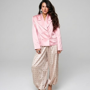 Puede incluir: Un blazer rosa claro de doble botonadura combinado con pantalones beige con lunares. El blazer tiene solapas con muescas y dos filas de botones. Los pantalones son de pernera ancha con tobillo fruncido. El conjunto es de material brillante.