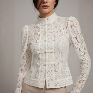 Puede incluir: Blusa de encaje color marfil con cuello mao y mangas largas. La blusa presenta cierres de rana decorativos en la parte delantera y un delicado estampado floral. Las mangas tienen un ligero abullonado en los hombros.