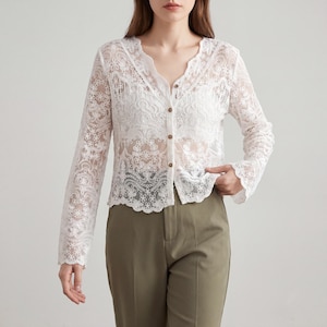 Könnte beinhalten: Weißer Spitzen-Cardigan mit langen Ärmeln, einem wellenförmigen Ausschnitt und Knopfverschluss. Das zarte Blumenmuster der Spitze ist deutlich sichtbar. Der Cardigan wird mit einer olivgrünen Hose kombiniert.