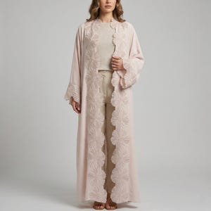 Puede incluir: Abaya abierta larga de color rosa claro con bordados florales en la parte delantera y las mangas. La abaya se lleva sobre un top y un pantalón beiges. El diseño floral se repite en las mangas y a lo largo de los bordes delanteros.