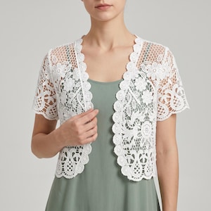 Op de afbeelding: Witte kanten bolero met korte mouwen en een bloemenpatroon. De jas heeft een geschulpte rand en wordt over een groene jurk gedragen. Het delicate kanten ontwerp is zichtbaar over het hele kledingstuk.