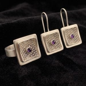 May include: A silver jewelry set consisting of a ring and a pair of drop earrings. Each piece features a square frame with a delicate silver filigree mesh center and a small, round, purple amethyst gemstone set in the middle.
