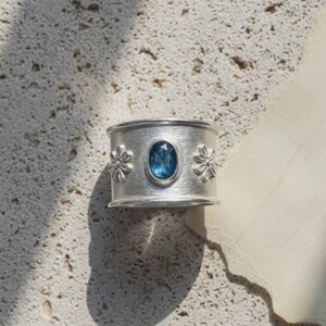 Pode incluir: Um anel de banda larga em prata com uma pedra preciosa azul de corte oval ao centro. A superfície do anel tem um acabamento mate escovado e é decorada com dois pequenos motivos florais em relevo, um de cada lado da pedra central.