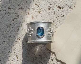 Könnte beinhalten: Ein breiter Silberring mit einem zentralen, oval geschliffenen blauen Edelstein. Die Metalloberfläche hat eine gebürstete, matte Textur. Auf beiden Seiten des zentralen Steins befindet sich ein kleines, erhabenes Blumenmotiv.