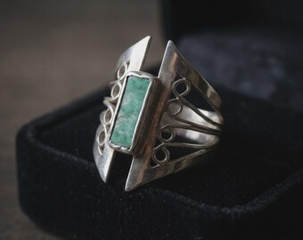 May include: A silver ring featuring a rectangular green stone set in the center. The band has a wide, open design with decorative wire scrollwork on both sides, creating a distinct geometric look with a polished metallic finish and natural stone.