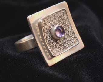 May include: A silver ring featuring a square-shaped face with a brushed metal finish. The center of the square contains a raised, intricate wire mesh panel with a small, round, purple gemstone set in a smooth silver bezel at its very center.