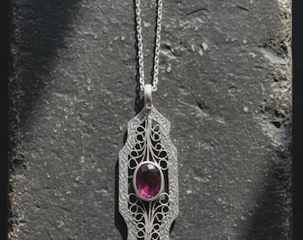 Op de afbeelding: Een zilveren hangerketting met een langwerpig, getextureerd metalen frame met ingewikkeld filigraanwerk en een centrale ovaalvormige, diep magenta edelsteen. De hanger hangt aan een fijne zilveren ketting.