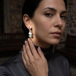 May include: A pair of silver-toned drop earrings featuring a vertical row of small, rectangular, iridescent white mother-of-pearl inlays. The earrings have a textured, metallic finish and a slender, elongated design that hangs from the earlobe.