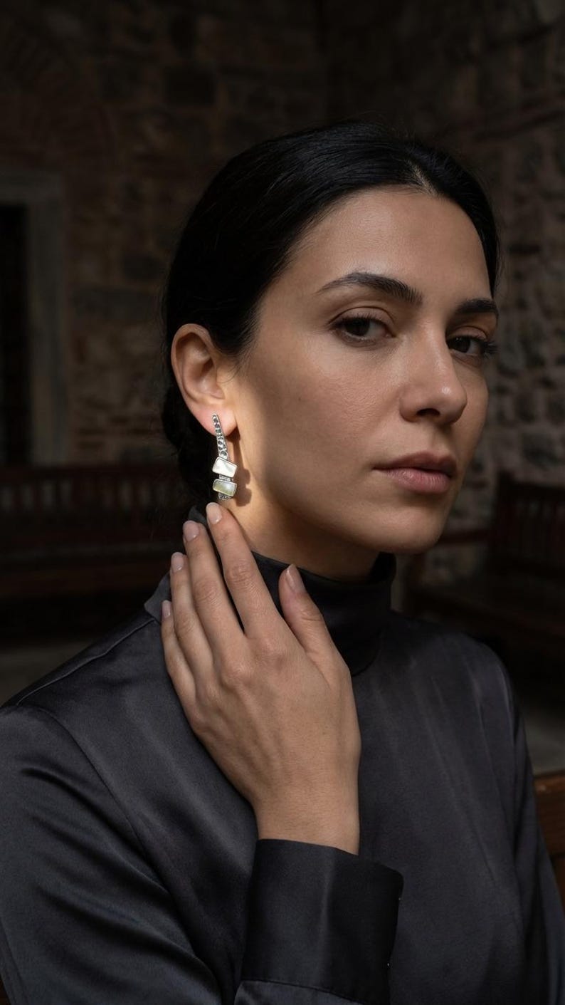May include: A pair of silver-toned drop earrings featuring a vertical row of small, rectangular, iridescent white mother-of-pearl inlays. The earrings have a textured, metallic finish and a slender, elongated design that hangs from the earlobe.
