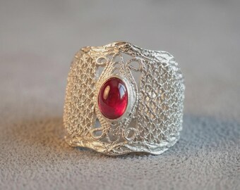Puede incluir: Un anillo ancho de plata con un intrincado patrón de malla texturizada en la banda. El centro del anillo está adornado con una piedra preciosa roja oscura, lisa y ovalada, engastada en un bisel de plata pulida y rodeada de filigrana.