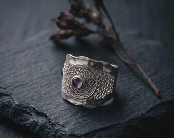 Puede incluir: Un anillo ancho de plata con acabado martillado y un panel central circular de filigrana. El centro del diseño de filigrana está engastado con una pequeña piedra preciosa redonda de color púrpura pulida. El aro tiene un borde irregular.