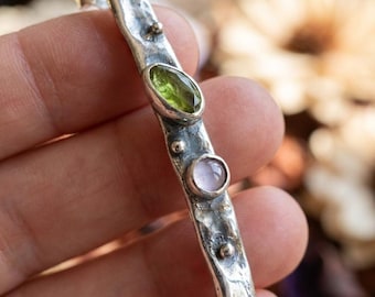 Könnte beinhalten: Eine silberne Stab-Brosche mit texturierter, gehämmerter Oberfläche, besetzt mit einem marquise-geschliffenen grünen Peridot und einem runden, blassrosa Cabochon, akzentuiert durch drei kleine, in die Metalloberfläche eingelassene Silberkugeln.