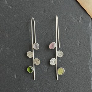 Puede incluir: Un par de pendientes colgantes de plata con un tallo de alambre vertical largo. Cada pendiente está adornado con tres elementos circulares: dos discos de plata texturizados y una piedra preciosa redonda y lisa en tonos verde y rosa.